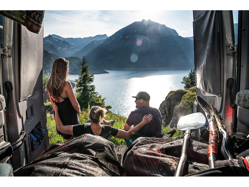 3 people sitting at the back of their Winnebago Revel looking out over a lake with mountains in the background.