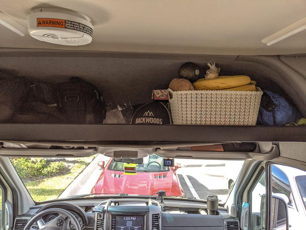 Overhead shelf above cab with white basket full of fruit, a baseball cap, a game and other supplies