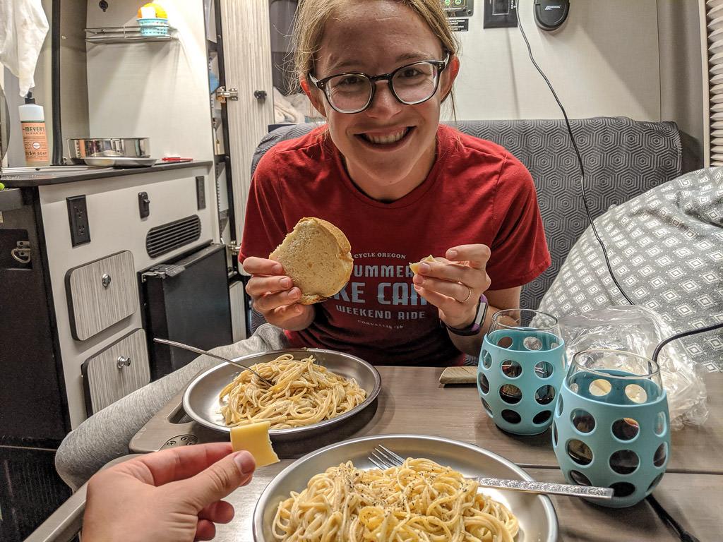 Kelly smiling at camera holding a piece of bread. Two plates of spaghetti and two glasses on the table in front of her. David's hand is in the picture holding a piece of cheese.