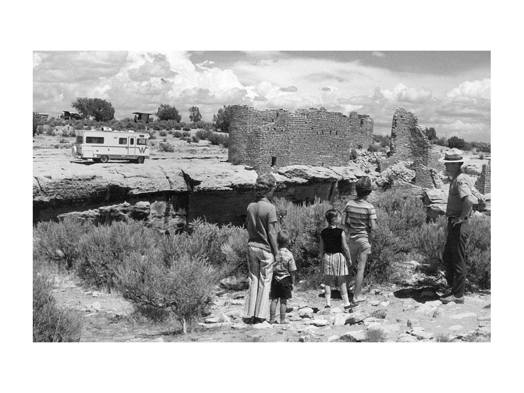 Black and white photo of park ranger leading a family of four through park. Old Winnebago Brave in the distance