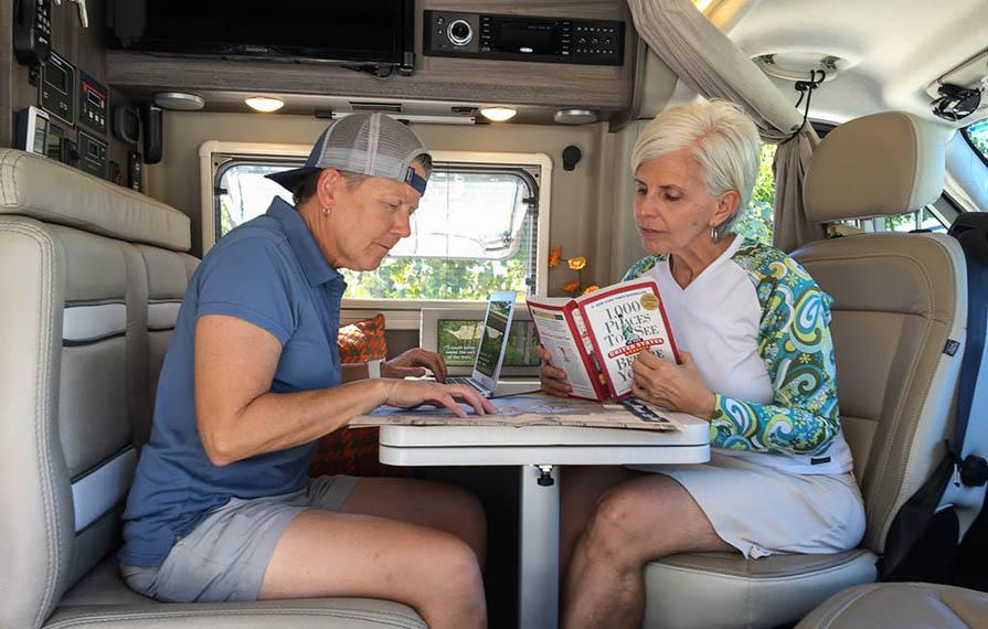 Noel and Chris sitting at dinette planning travels. Noel is looking at a map with laptop open and Chris is reading a travel destination book