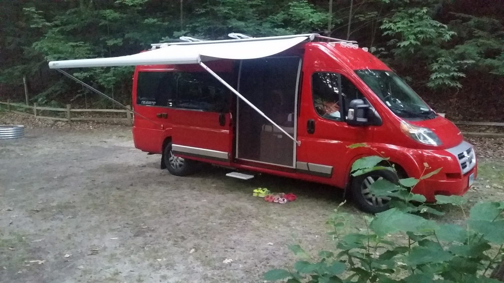 Winnebago Travato with awning out in tree surrounded campsite.