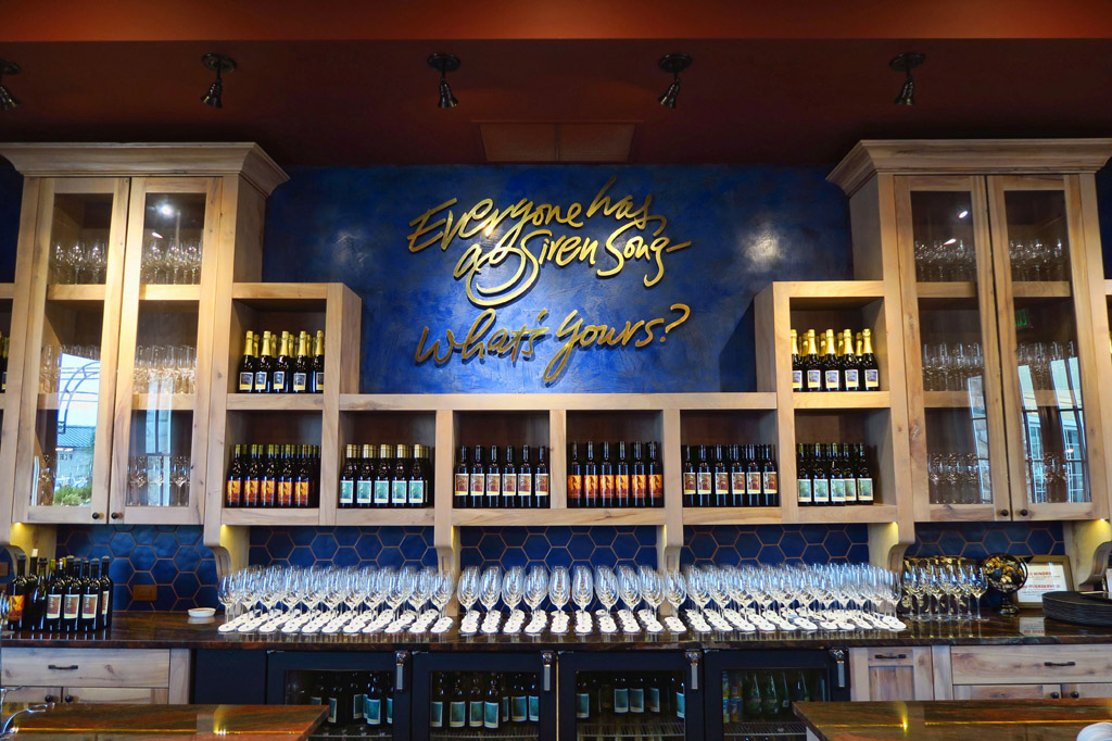 Shelving lining wall filled with wine bottle and words on the wall that say, "Everyone has a siren song. What's Yours?"