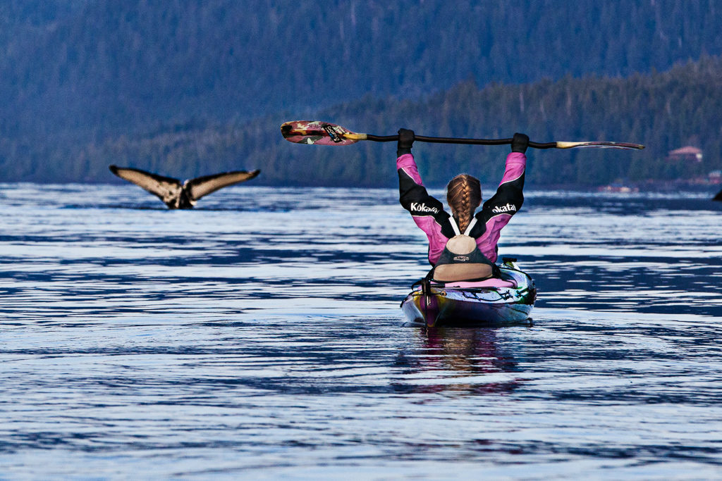 Abby on the water in her kayak with arms raised in victory as a whale tale dips back into the water ahead.