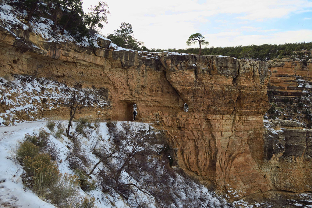 Hiker going though a small tunnel in the rock to continue on the snow covered path.