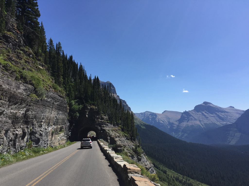 Cars traveling along mountain side road about to pass under a short tunnel with tree filled valley below.