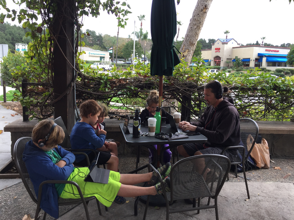 Craig working on a computer and four kids watching tablets at a table outside Starbucks.