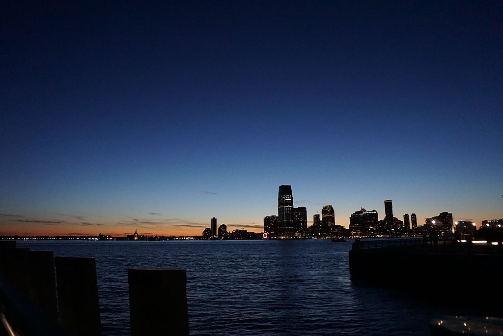 Downtown NYC with buildings lit up across the water against the setting sun.