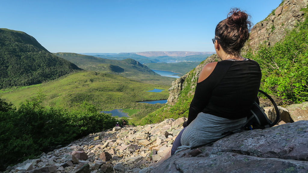 Woman resting on rock looking out over green lush valley with water running below.