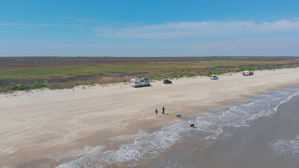 Winnebago Navion and other cars parked along the beach with couple and dogs walking along the water