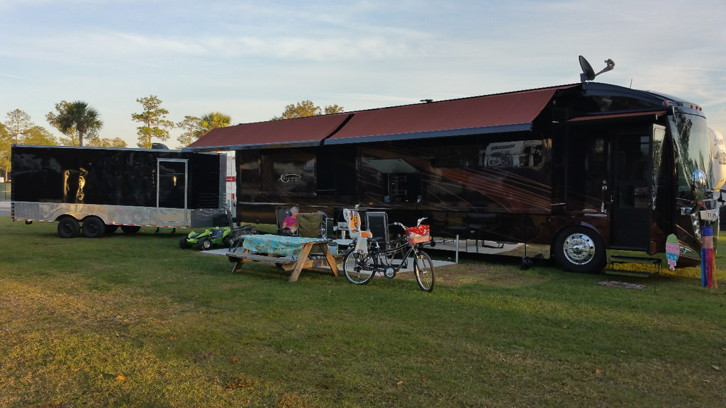 Adventurer with awnings extended parked in grass. Black enclosed trailer is parked behind it.