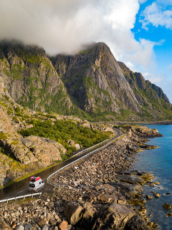 Winnebago Revel parked on a small turn off along the water with mountainside to the left with clouds looming overhead.